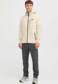 Beige fleece hoodie med sort lynlås og brystlomme, parret med grå sweatpants. Modellen står oprejst, iført grå og hvide sneakers.