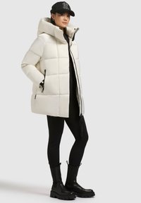 Fehér puffernadrág kapucnival, cipzáras zárással, oldalzsebekkel és steppelt dizájnnal. Fekete leggings-szel és vastag fekete csizmával viselve.