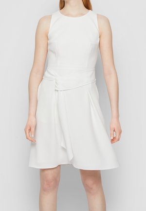 Femme portant une robe blanche sans manches avec un corsage ajusté et un détail drapé à la taille, debout devant un fond clair uni.