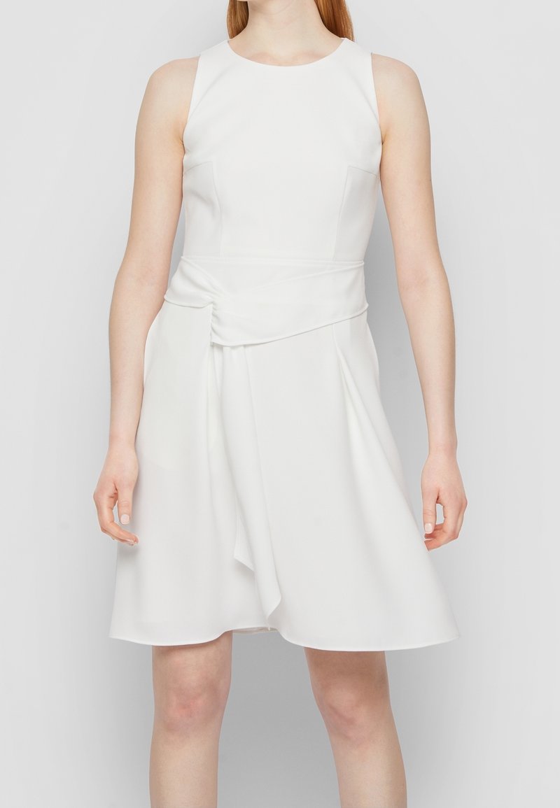 Femme portant une robe blanche sans manches avec un corsage ajusté et un détail drapé à la taille, debout devant un fond clair uni.