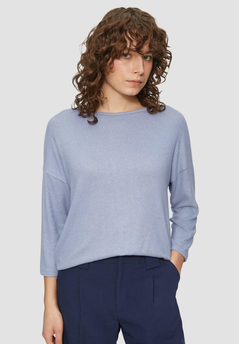 Vrouw met krullend bruin haar, gekleed in een lichtblauwe lange mouwen trui en donkerblauwe highwaisted broek, staande met één hand in de zak.