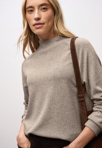 Pull à col roulé en tricot gris clair dans un tissu doux. Le mannequin tient un sac à bandoulière en cuir marron, se tenant devant un fond neutre.
