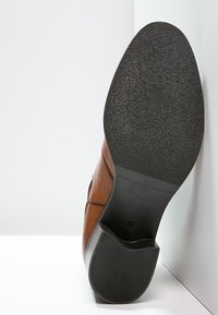 Scarpa di pelle marrone con suola in gomma nera testurizzata, caratterizzata da una punta arrotondata e cuciture visibili lungo il bordo superiore.