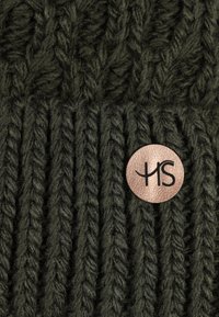Knit beanie in dark green with a textured pattern. Features a ribbed band and a round metallic tag with "HS" in black lettering.