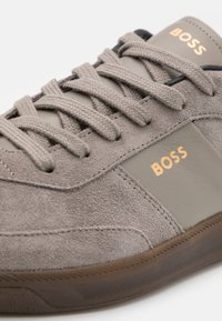 Šedá semišová teniska s béžovými koženými detaily, plochými tkaničkami a hnědou gumovou podrážkou. Na straně se zlatým logem „BOSS“.