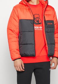 Vadderad jacka med orange och svart färgblockdesign, dragkedjestängning, justerbar huva och elastiska ärmslut. Bärs över en röd sweatshirt.