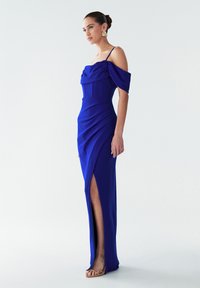 Žena v královsky modré off-shoulder večerní šaty s bočním rozparkem, tenkými ramínky, nařasenými detaily a béžovými otevřenými lodičkami, stojící na bílém pozadí.