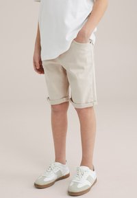 Beige džinsa šorti ar ruļļveida malām, sānu kabatām un gludu tekstūru, apvienoti ar baltām un pelēkām sporta apaviem.