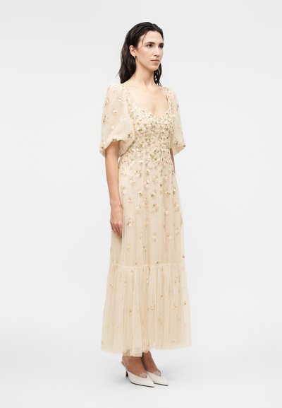 Femme portant une longue robe beige transparente avec broderies florales et manches bouffantes, associée à des escarpins beiges à bouts pointus, debout devant un fond uni.