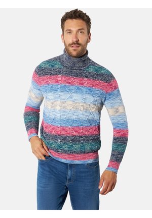 Mann trägt einen mehrfarbigen gestreiften Rollkragenpullover und blaue Jeans und steht mit einer Hand in der Hosentasche.