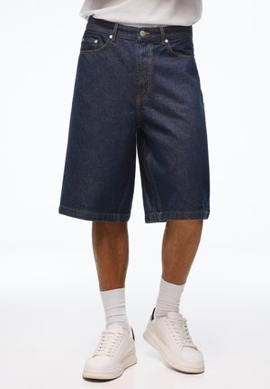 Férfi sötétkék farmer rövidnadrágban, fehér zokniban és fehér sportcipőben, sima fehér háttér előtt áll.