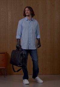 Lys blå skjorte med knapper, mørke jeans, svart lærveske, hvite sneakers og svarte hansker. Trebakgrunn, avslappet stående posisjon.