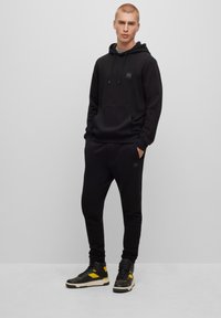 Felpa nera con cappuccio e pantaloni jogger coordinati realizzati in tessuto morbido. La felpa presenta una tasca frontale e lacci. Il modello indossa sneakers nere e gialle.