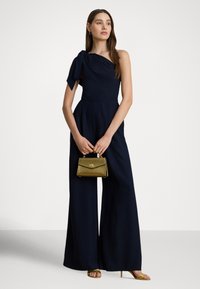 Granatowy kombinezon na jedno ramię z kokardą, o szerokich nogawkach, zestawiony ze złotą metaliczną torebką i złotymi sandałami na paski.