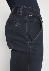 Mörka denimjeans med en slät textur, med en flerficksdesign och en liten sydd logotyp i rött, vitt och blått på bakfickan.
