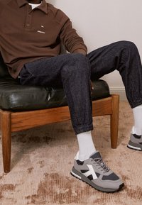 Polo a maniche lunghe marrone, joggers in denim scuro con polsini elastici e sneakers grigie con dettagli bianchi. Seduto su una sedia in pelle nera.