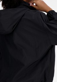 Veste coupe-vent noire, légère, avec un col montant, des poignets élastiques et une texture lisse légèrement froissée.
