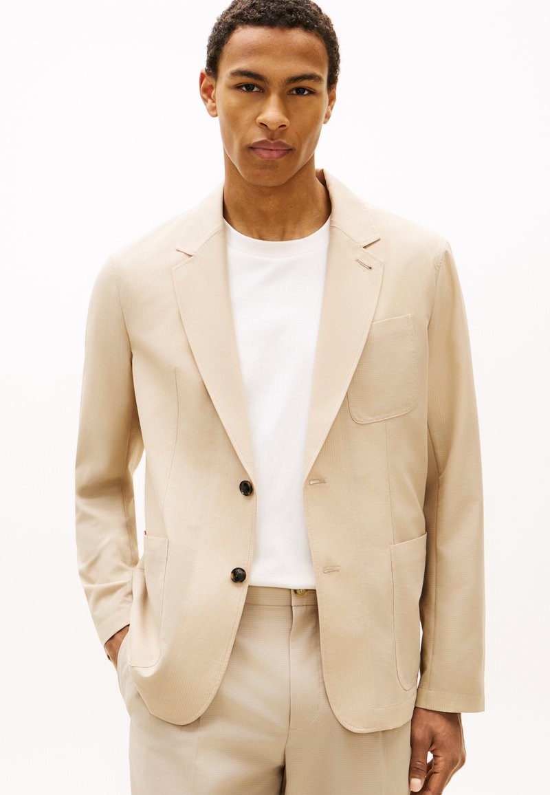 Νέος άνδρας που φοράει μπεζ σακάκι και παντελόνι που ταιριάζουν με λευκό t-shirt, στέκεται με το ένα χέρι στην τσέπη μπροστά σε απλό φόντο.