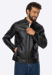 Man glimlachend terwijl hij zijn zwarte leren jas dichtritst, met één hand in zijn zak en een blauwe spijkerbroek aan.