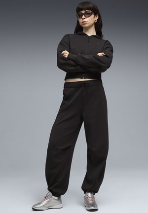 Kobieta w czarnej bluzie rozpinanej na zamek i dopasowanych spodniach dresowych, srebrnych trampkach i ciemnych okularach przeciwsłonecznych, stojąca z założonymi rękami na tle szarego tła.
