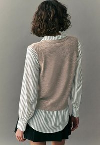 Beżowy sweter w serek noszony na białej koszuli w paski z długimi rękawami. Warstwowano to z czarną plisowaną spódnicą. Gładka tekstura, codzienny fason.