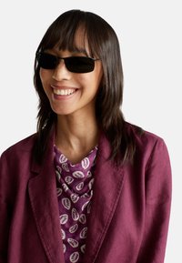 Des lunettes de soleil avec des montures noires et des verres foncés. La personne qui les porte a les cheveux noirs raides et est habillée d'une veste violette et d'un haut à motifs.