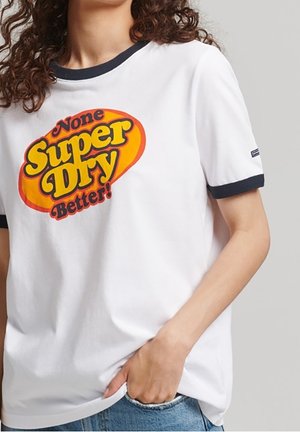 Femme portant un t-shirt blanc avec col et manches bordés de noir, arborant un logo orange et jaune "None Super Dry Better!", main dans la poche du jean.