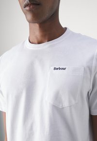 Biała bawełniana koszulka z okrągłym dekoltem, kieszonką na lewej piersi oraz granatowym haftowanym logo "Barbour" na kieszeni. Miękka tekstura, swobodny styl.