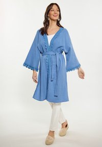 Kimono din bumbac albastru cu cordon, având garnituri din dantelă ondulată pe mâneci și tiv, purtat peste un top alb și asortat cu pantaloni albi.