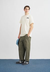 Μπλούζα από βαμβάκι σε κρεμ χρώμα με λογότυπο, φαρδιά χακί παντελόνια cargo και μαύρα αθλητικά παπούτσια με λευκές λεπτομέρειες. Κρατά ένα μπλε τζιν αντικείμενο.