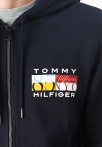 Sweat-shirt zippé marin avec un logo géométrique coloré et le texte "TOMMY 85 NY" et "HILFIGER" en blanc sur la poitrine. Matière douce en mélange de coton.