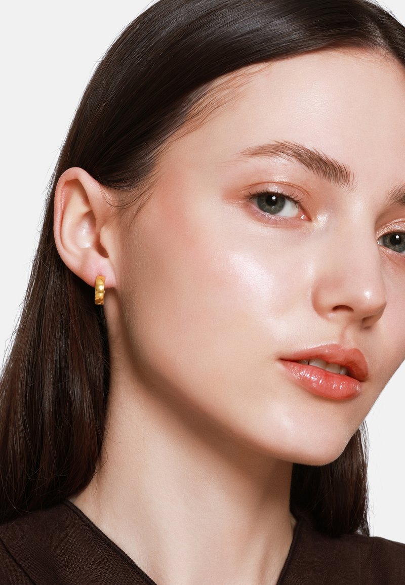 Gouden hoepel oorbel met een gestructureerd oppervlak, gedragen door een model met een gladde huid en neutrale make-up, met lang donker haar en een bruine top.