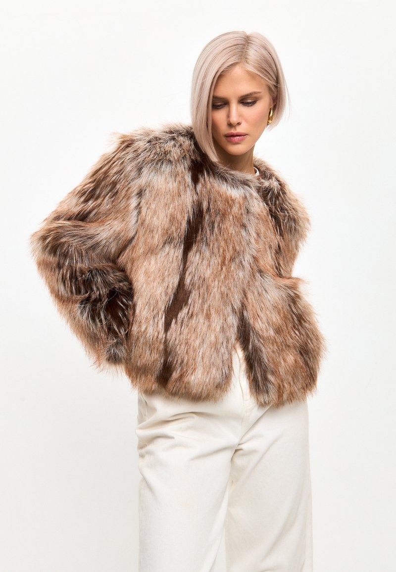 Chaqueta corta de piel sintética marrón con una textura suave, que presenta un cuello alto y un corte holgado. Combinada con pantalones de color crema.