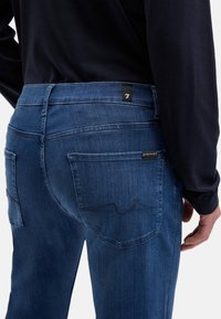 Ciemnoniebieskie dżinsy z dopasowanym krojem, z widocznymi przeszyciami, dwoma tylnymi kieszeniami i małą firmową metką na pasku talii.