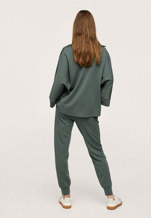 Kobieta o długich brązowych włosach, ubrana w luźną ciemnozieloną bluzę i dopasowane spodnie dresowe, stoi tyłem, ma na nogach białe sneakersy z gumowymi podeszwami.