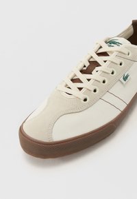 Zapatillas de lona con parte superior beige y crema, ojetes y cordones marrones, suela de goma marrón texturizada, logotipo en el lateral.