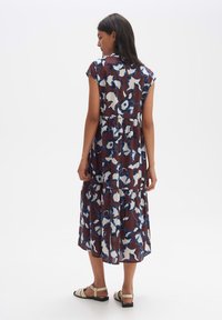 Ärmelloses, gestuftes Kleid aus braunem Stoff mit blau-weißen Blumenmustern, leichtes Material, knielang, kombiniert mit flachen Sandalen.