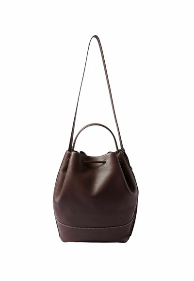 Bolso de cuero marrón tipo cubo con base redonda, cierre de cordón y correa larga. Presenta una textura suave y un detalle de costura blanca en contraste.