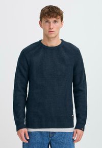 Dark navy knitted sweater with a ribbed texture, long sleeves, and a round neckline. Features a subtle logo tag near the hem.