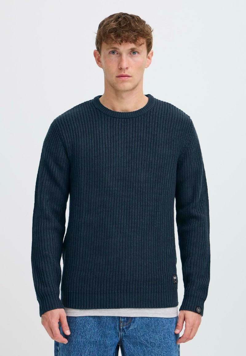 Dark navy knitted sweater with a ribbed texture, long sleeves, and a round neckline. Features a subtle logo tag near the hem.