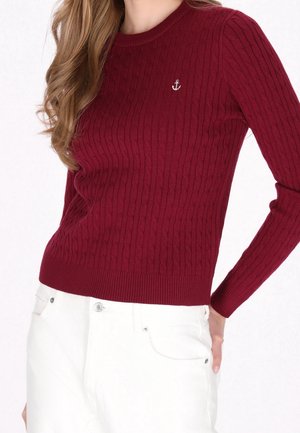 Femme portant un pull en maille torsadée bordeaux avec un petit logo d'ancre sur la poitrine et un pantalon blanc, posant avec la main sur la hanche.