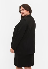 Blazer nero su misura con maniche lunghe, revers, e una vestibilità rilassata, abbinato a una gonna nera coordinata, con dettagli minimi.