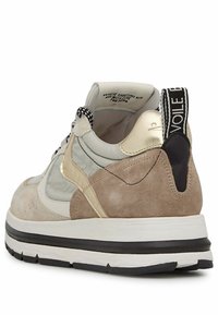 Zapatilla con parte superior de ante beige, acentos metálicos dorados, lengüeta trasera negra, entresuela blanca y patrón texturizado en el panel lateral.