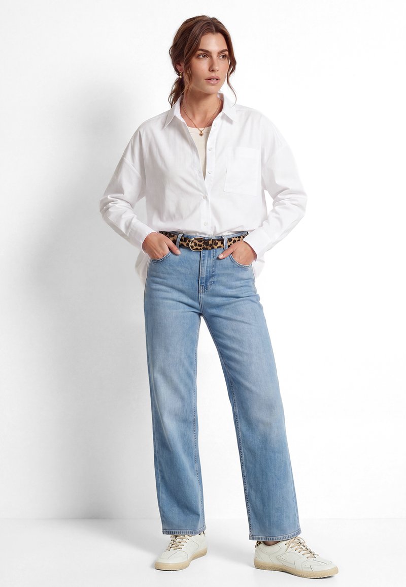 Femme debout, les mains dans les poches, portant une chemise blanche à boutons, un jean bleu clair, une ceinture imprimé léopard et des baskets blanches sur fond blanc.