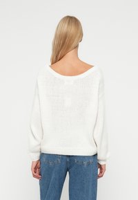 Biały sweter dziergany z wycięciem z tyłu, w oversize'owym kroju i żebrowanym dołem, zestawiony z jasnoniebieskimi dżinsami z wysokim stanem.