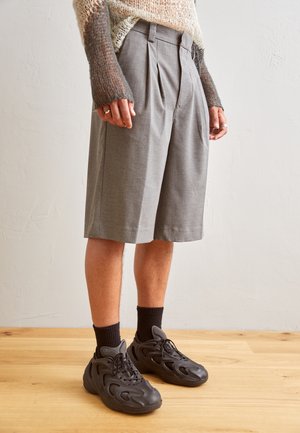 Person wearing gray pleated knee-length shorts, black socks, chunky black sneakers, and a loose knit sweater with gradient brown sleeves.