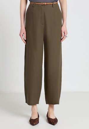 Femme portant un pantalon large taille haute couleur vert olive avec une ceinture marron tressée et des ballerines pointues imprimé léopard, debout sur un fond uni.