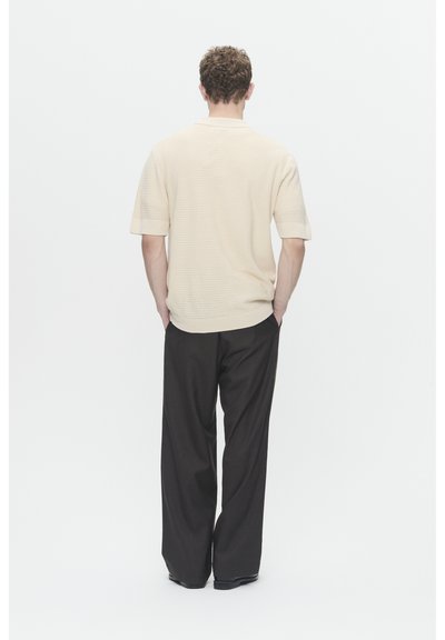 Chemise en tricot beige à manches courtes avec un motif texturé, associée à un pantalon large noir. Le mannequin se tient avec les mains dans les poches.