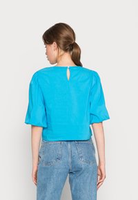Blusa azul de manga curta, cropped, em tecido leve, com mangas bufantes e uma pequena abertura em forma de olho de fechadura nas costas, acabada com um botão.