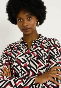 Chemise à boutons avec un motif géométrique en noir, rouge et crème, à manches longues et col classique. Texture de tissu lisse.